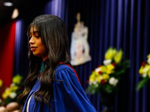 A woman in a blue academic robe smiles while crossing the stage during a university graduation ceremony.