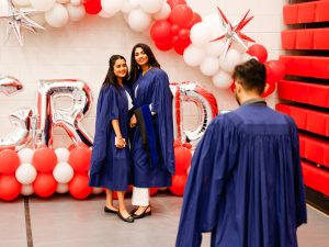 A person in a blue academic robes takes a photo of two young women also wearing blue robes in front of a red and white balloon arch before a university graduation ceremony.