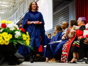 A young woman in a blue academic gown smiles while walking across the stage during a university graduation ceremony. A group of people in academic regalia watch her while seated on the stage.