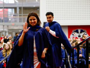 A young woman in a blue academic gown waves while leaving the stage during a university graduation ceremony. Another new graduate is walking down the stairs behind her.