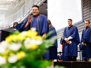 A young man in a blue academic gown waves to the audience while walking across the stage during a university graduation ceremony. Other graduating students wait in line behind him.