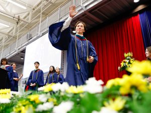 A young man in a blue academic gown waves to the audience while walking across the stage during a university graduation ceremony.