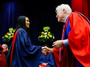 A young woman in a blue academic gown shakes hands with a professor in red academic regalia on stage during a university graduation ceremony.