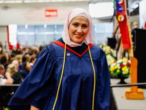 A women wearing a hijab and blue academic gown smiles while leaving the stage during a university graduation ceremony.