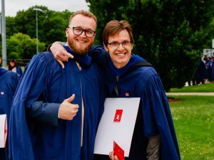 Two new university graduates pose for a photo while wearing blue academic gowns and holding their diplomas.