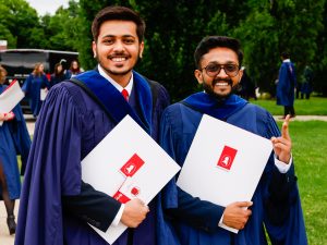 Two new university graduates pose for a photo while wearing blue academic gowns and holding their diplomas.