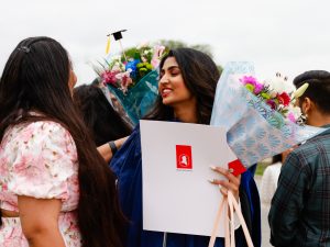 A young woman in a blue academic gown holds flowers and a diploma while hugging a loved one after a university graduation ceremony.