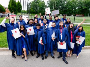 A large group of new university graduates in blue academic gowns hold their diplomas and smile for a photo outside a graduation ceremony.