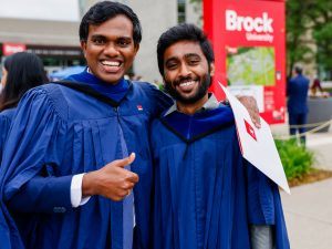 Two new university graduates pose for a photo while wearing blue academic gowns and holding their diplomas.