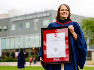 A new university graduate poses for a photo while wearing a blue academic gown and holding a framed diploma.