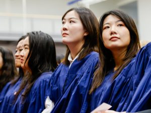 Three young women in blue academic robes sit in the audience during a university graduation ceremony.