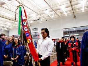 A man carrying a ceremonial staff leads a group of people wearing academic regalia into a university graduation ceremony.