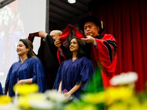 Two woman in a blue academic robes stands still while professors place academic hoods on their shoulders on stage during a university graduation ceremony.