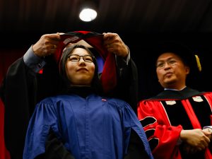 A woman in a blue academic robe stands still while someone places an academic hood on her shoulders and a professor in red academic regalia observes from beside her.