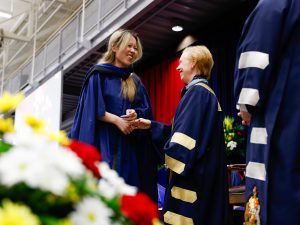 Two women in blue academic robes shakes hands on stage during a university graduation ceremony.