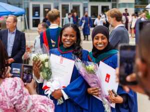 Two women in graduation gowns and holding university degrees pose for photos taken by people in the foreground with cellphones.