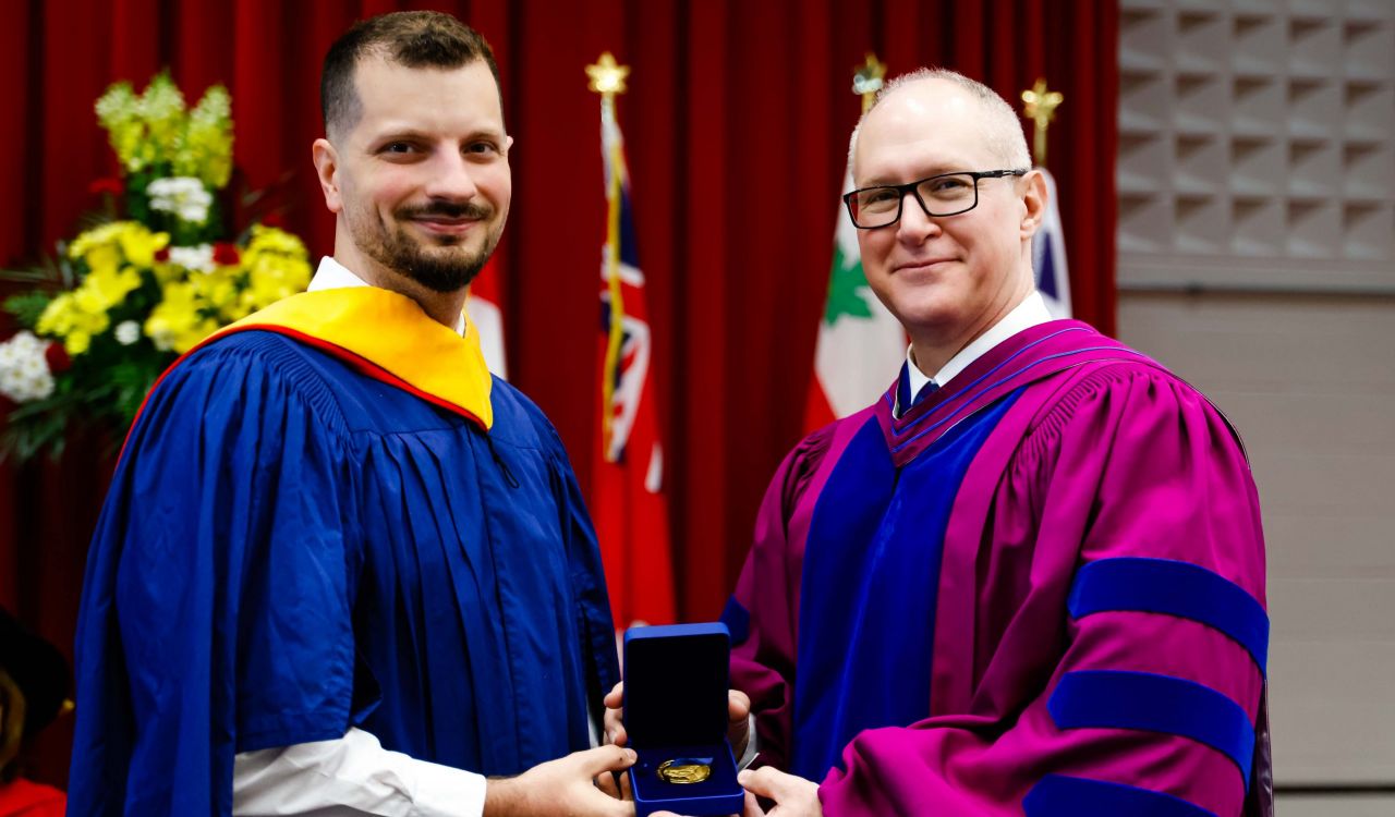 Two men in academic robes smile for a photo while holding a medal between them in a box.