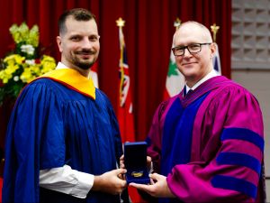 Two men in academic robes smile for a photo while holding a medal between them in a box.