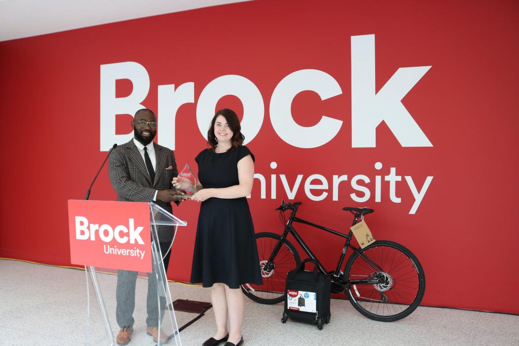 A man and woman stand behind a podium in a building atrium. The woman is holding a glass award.