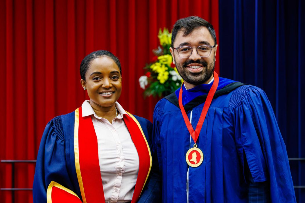 A woman wearing blue and red academic regalia poses for a photo with a man in a blue robe wearing a red and gold medal during a university graduation ceremony.