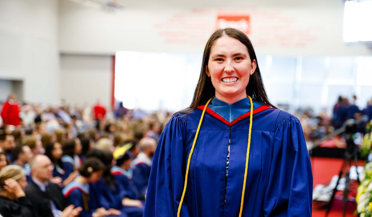 A woman in a graduation gown stands in front of a gymnasium filled with seated graduates.