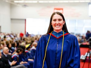 A woman in a graduation gown stands in front of a gymnasium filled with seated graduates.