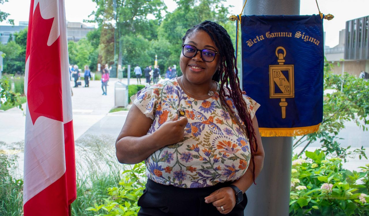 A woman stands in front of a Canadian flag and a blue flag representing the Beta Gamma Sigma organization.