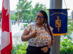 A woman stands in front of a Canadian flag and a blue flag representing the Beta Gamma Sigma organization.