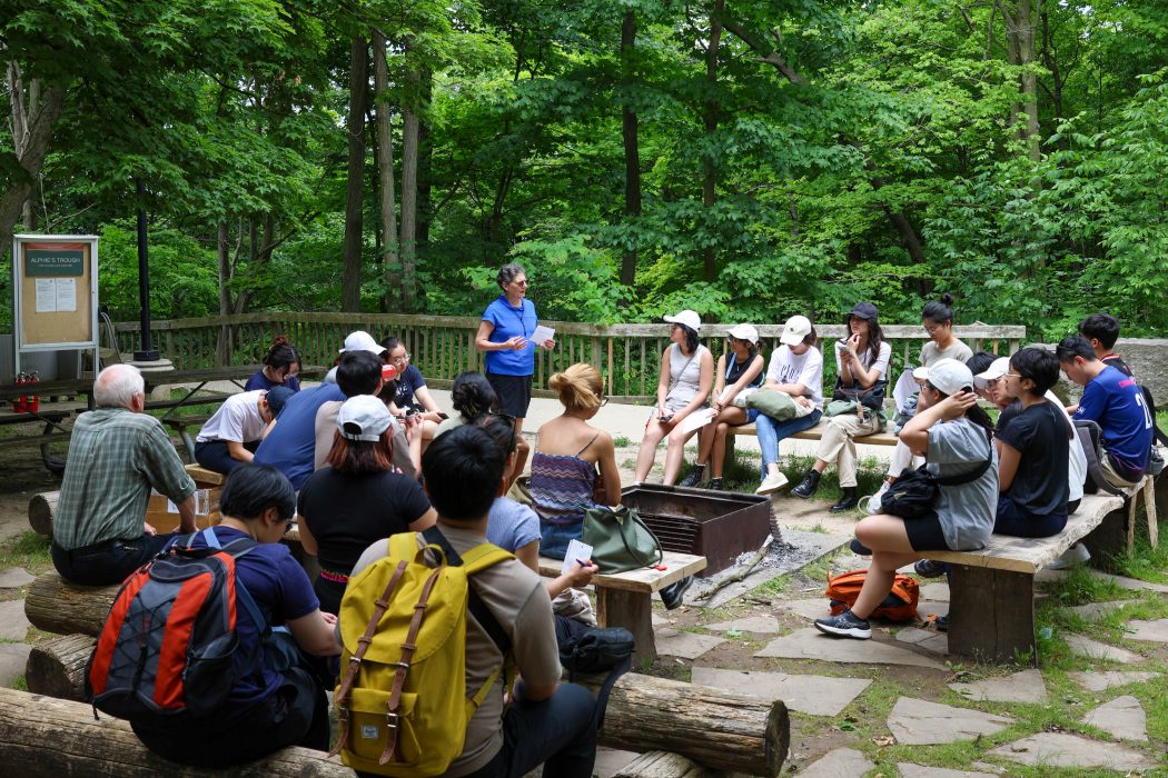 A woman speaks to a group of people sitting on benches outside around a fire pit among trees.