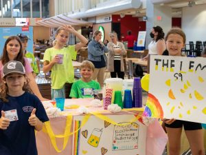 Six kids stand around a table with cups and a jug of lemonade. The girl on the far left holds a sign promoting their lemonade.