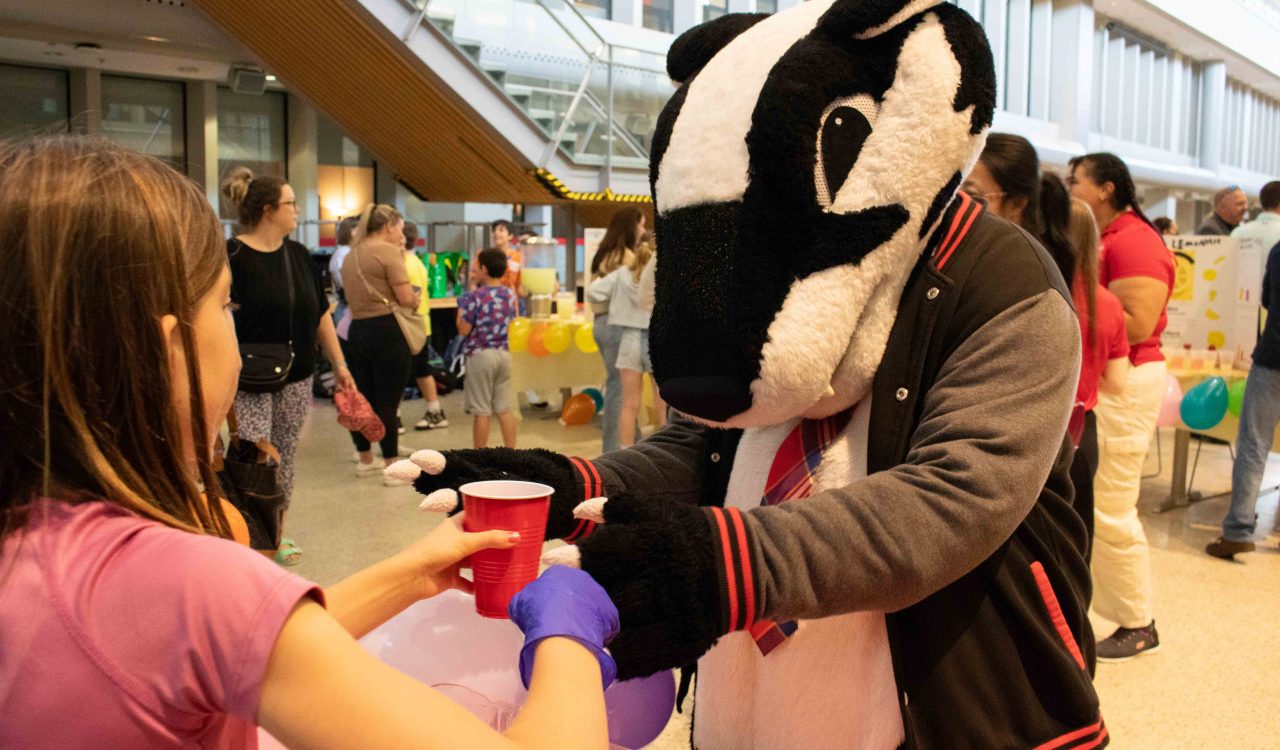 A mascot of a badger grabs a glass of lemonade from a fourth-grade student.