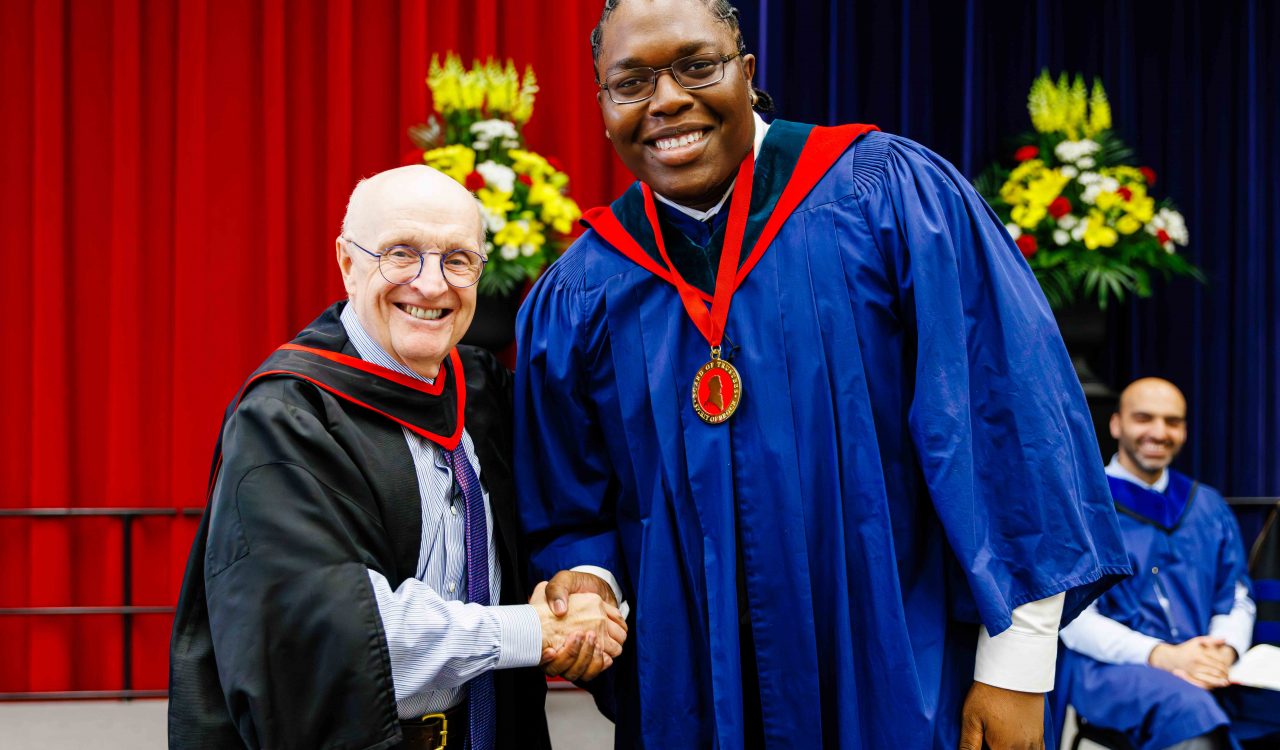 A man wearing black and red academic regalia shakes hands with a man in a blue robe wearing a red and gold medal during a university graduation ceremony.