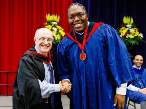 A man wearing black and red academic regalia shakes hands with a man in a blue robe wearing a red and gold medal during a university graduation ceremony.