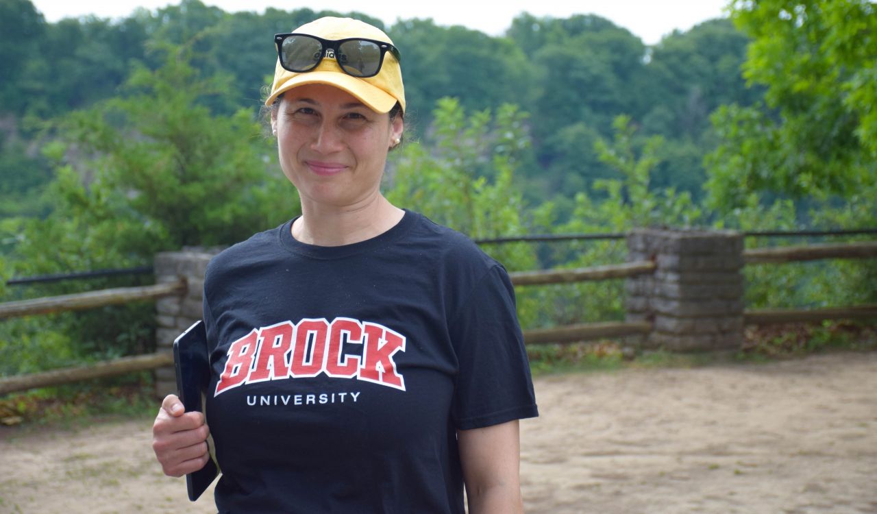 Jennifer Holzer, wearing a Brock University T-shirt and yellow baseball cap with sunglasses balanced on the brim, holds a tablet for survey data collection at the Niagara Glen.