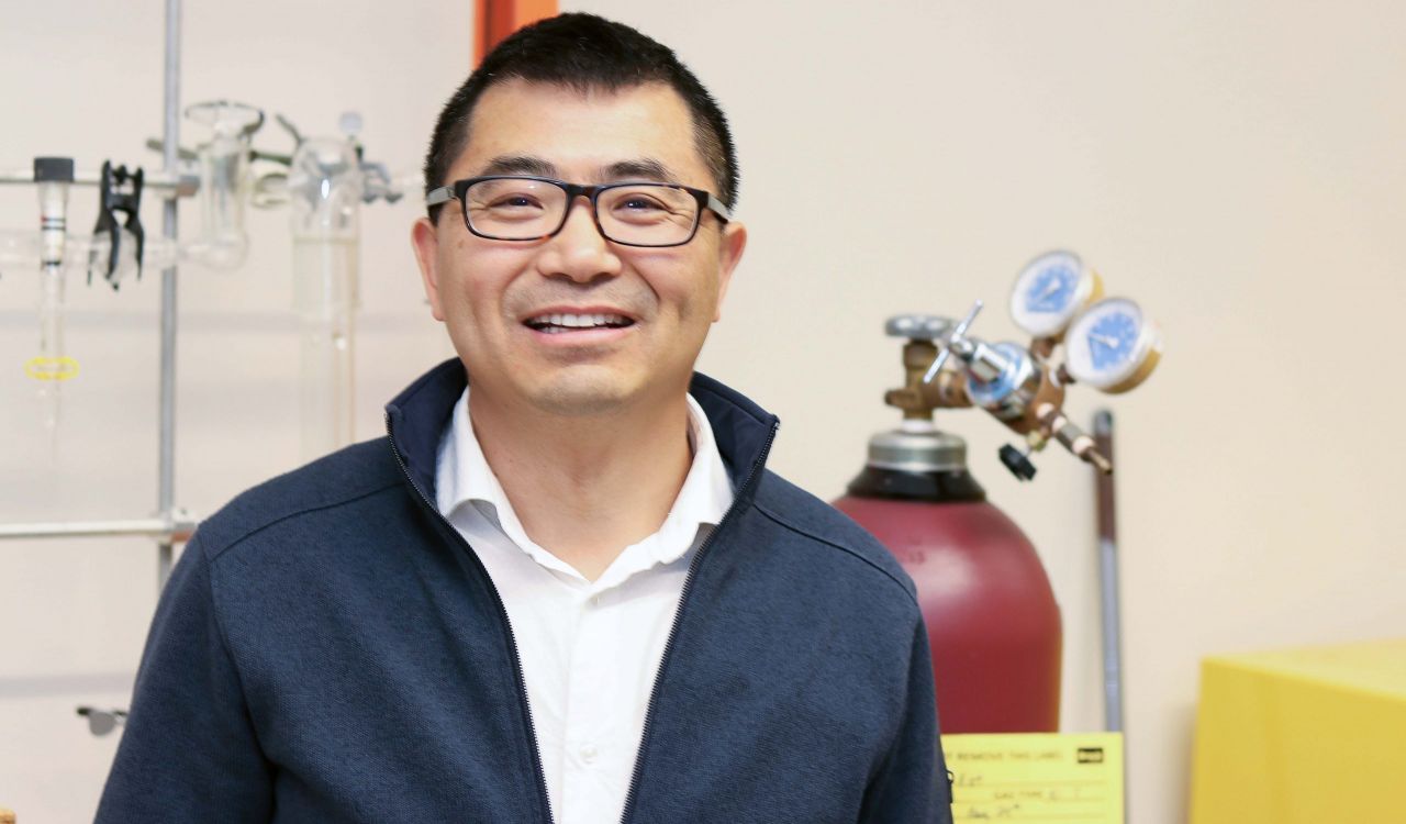 Close-up of Assistant Professor of Chemistry Jianbo Gao smiling, with lab equipment in the background.