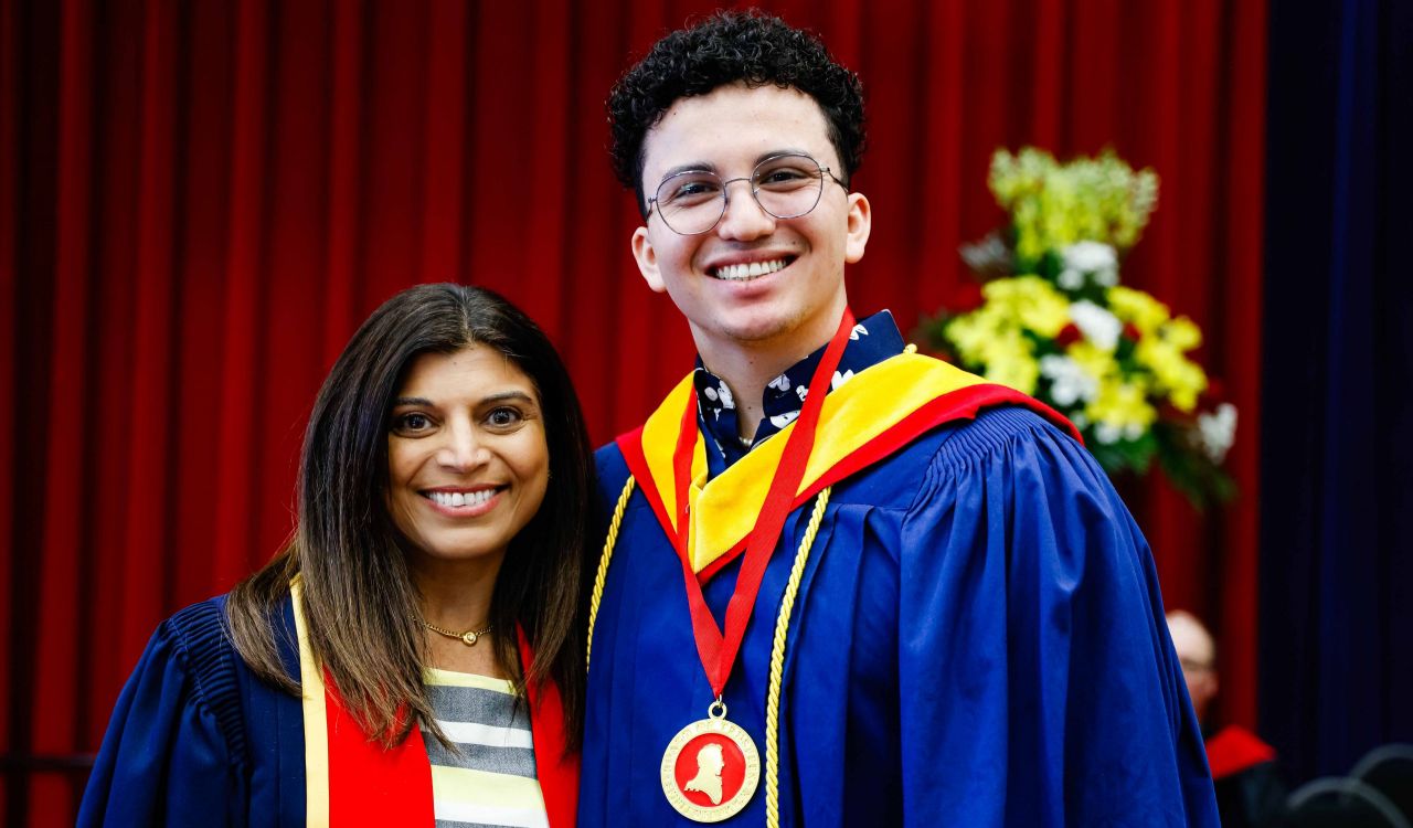 A young man wearing a blue academic robe and a medal poses for a photo next to a woman in academic regalia.