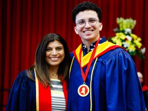 A young man wearing a blue academic robe and a medal poses for a photo next to a woman in academic regalia.