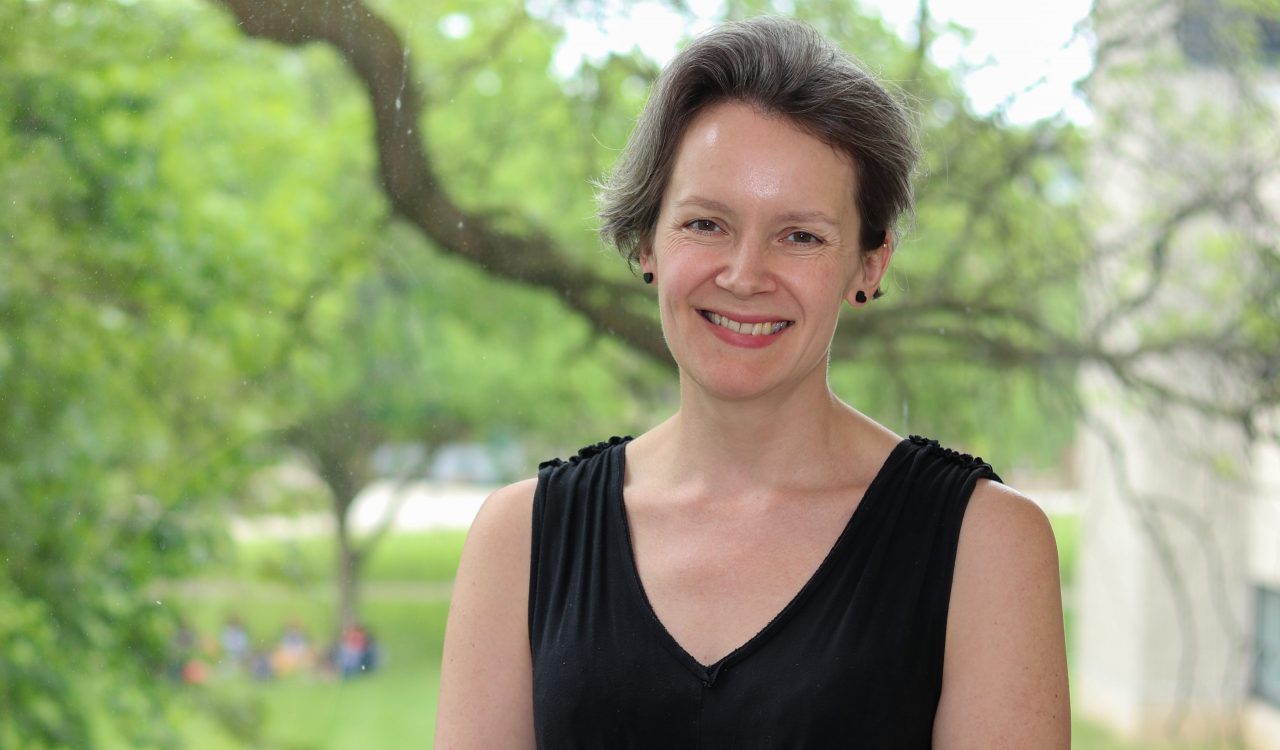 Waist-up view of Associate Professor of Psychology Karen Campbell standing in front of a window, with out-of-focus leaves and branches in the background.