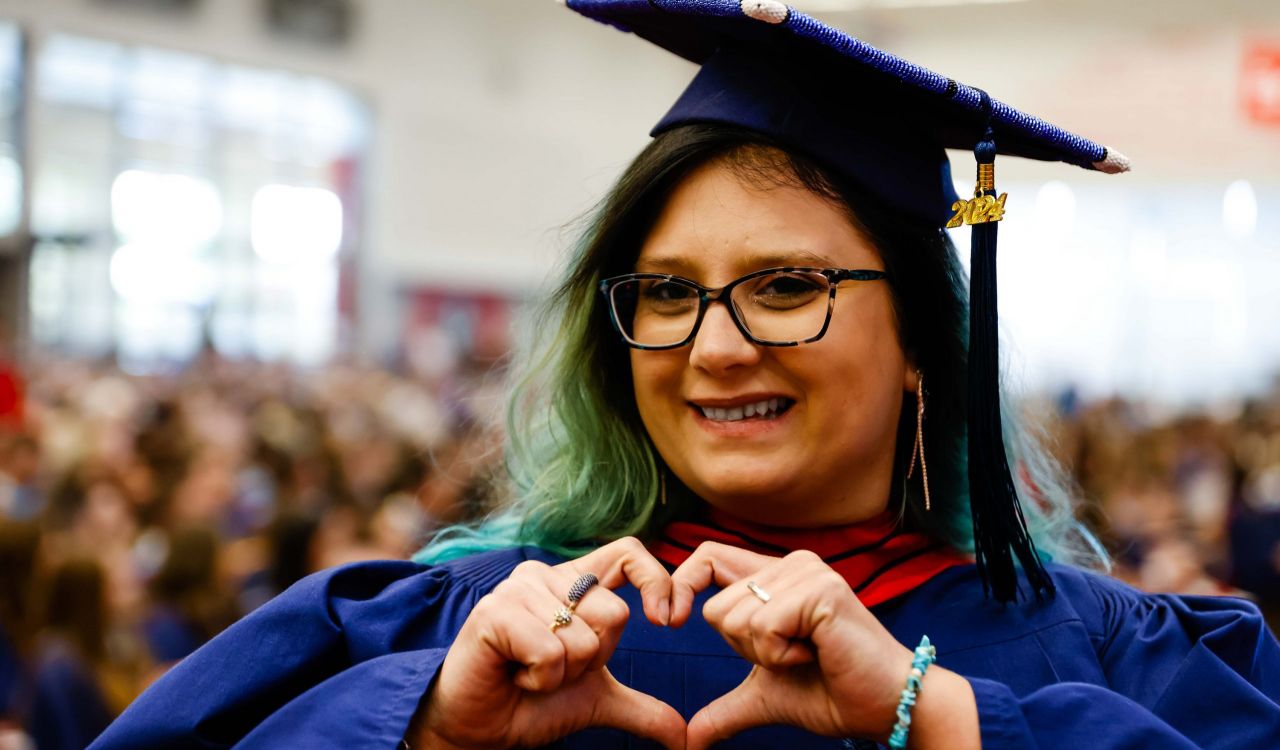 A woman wearing a graduation cap and gown makes a heart symbol with her hands.