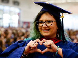 A woman wearing a graduation cap and gown makes a heart symbol with her hands.