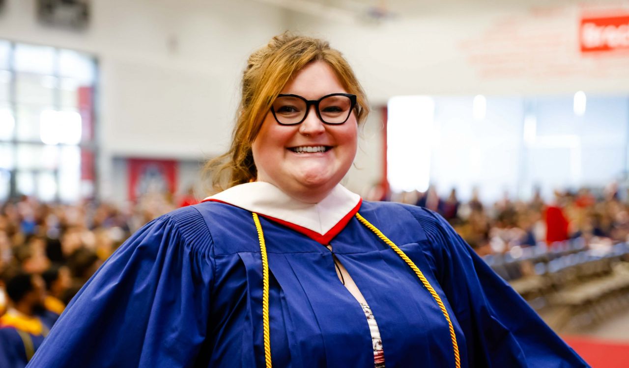 A woman in a blue academic robe stands in a sunny gymnasium during a university graduation ceremony. The audience and other graduates can be seen behind her.