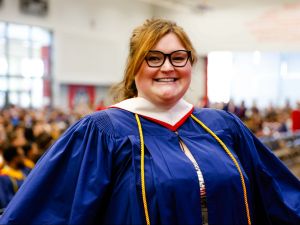 A woman in a blue academic robe stands in a sunny gymnasium during a university graduation ceremony. The audience and other graduates can be seen behind her.