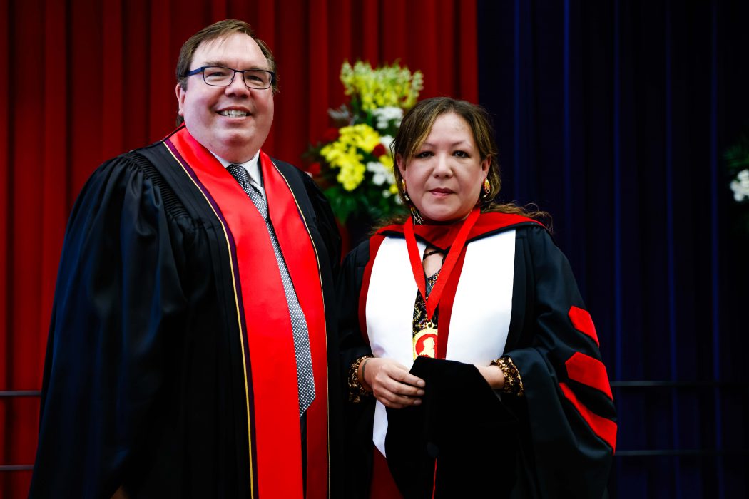 A man and a woman in academic robes pose for a photo on stage during a university graduation ceremony.