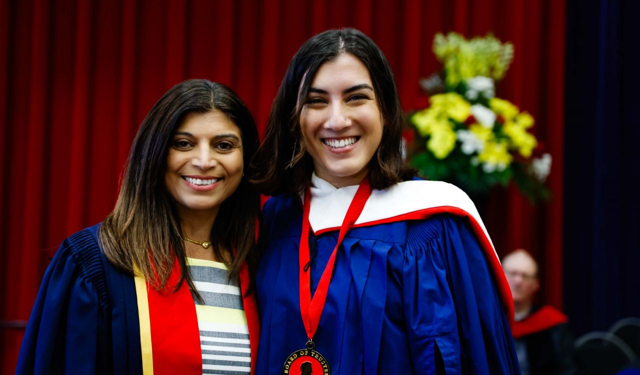 A young woman wearing a blue academic robe and a medal poses for a photo next to a woman in academic regalia.