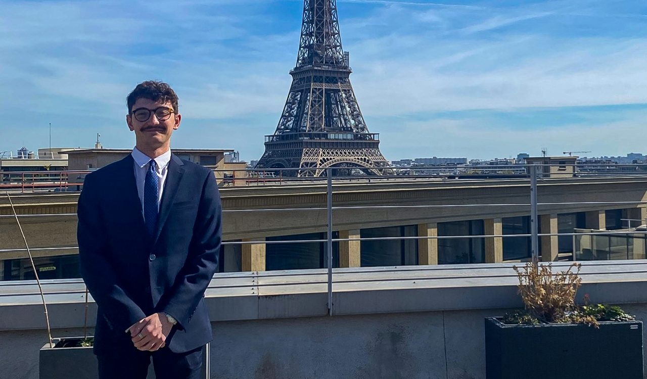 A man in a suit stands to the left side of the Eiffel Tower in Paris, France.