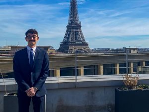 A man in a suit stands to the left side of the Eiffel Tower in Paris, France.