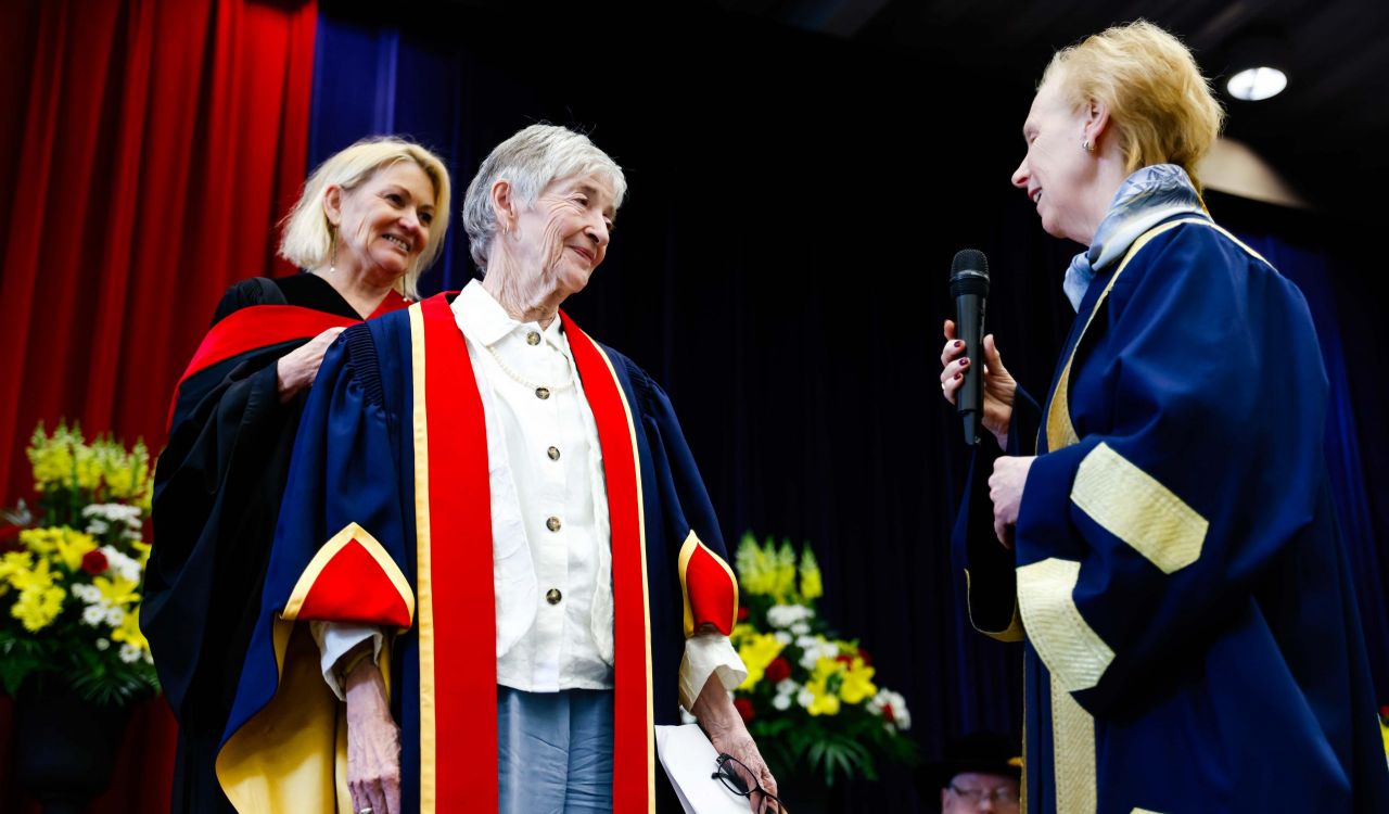 Three people in Convocation gowns stand together on a stage. One is holding a microphone.