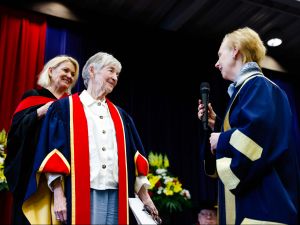 Three people in Convocation gowns stand together on a stage. One is holding a microphone.