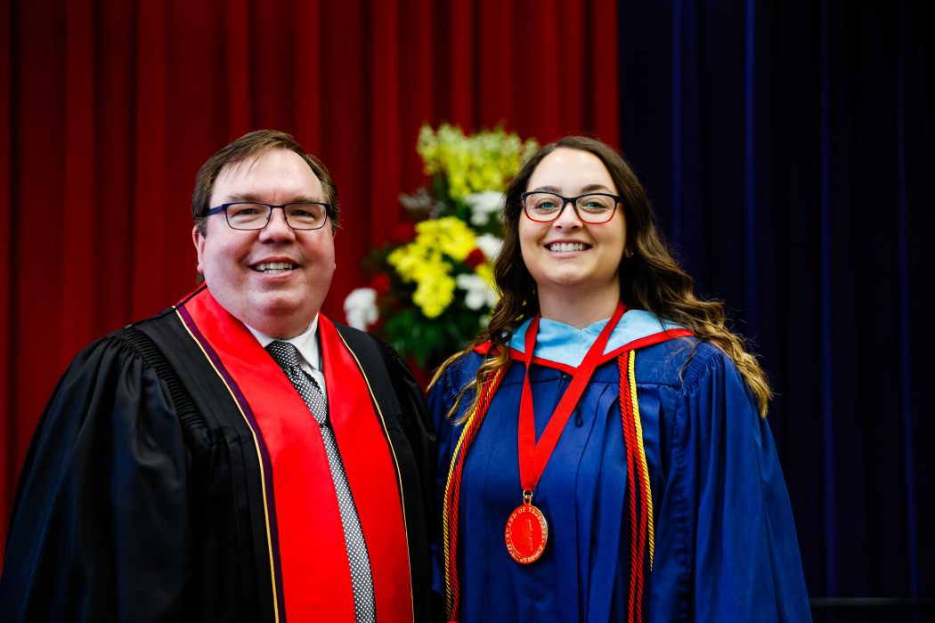 A young woman wearing a blue academic robe and a medal poses for a photo next to a man in academic regalia.