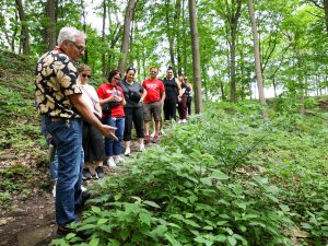 A man leads a group of 10 people on a nature hike in a wooded area, pointing to plants while talking.